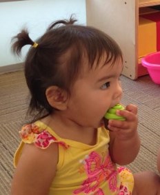 Bébé assis sur le plancher d’une salle de jeu, regardant loin de la caméra, mettant un petit jouet en plastique dans sa bouche.