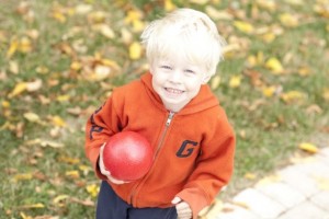 Jeune enfant à l’extérieur un jour d’automne devant de l’herbe avec des feuilles. Enfant tenant une grosse balle dans une main et se tenant debout, il regarde la caméra avec un grand sourire.