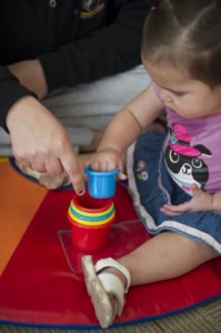 Bébé assis sur un tapis coloré et jouant avec des tasses colorées et empilées. Un adulte est assis et montre la pile de tasses.
