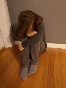 Sad looking child sitting on floor with head down on hands crossed at knees.