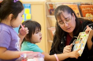 Two young children looking and pointing at a book held up to them by a sitting adult.
