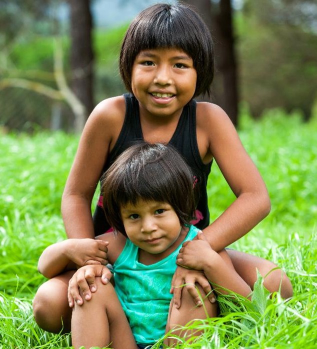 A young child sits in front of an older sibling in the grass.